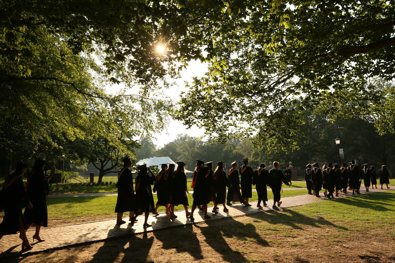 People in caps and gowns walk down a pathway in a wooded area