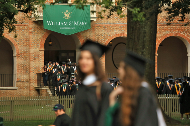 People in caps and gowns stream into a brick building with a William & Mary banner hanging on it.