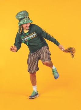 Phil Woodward strikes a dance move while wearing a green hat and holding a pom-pom