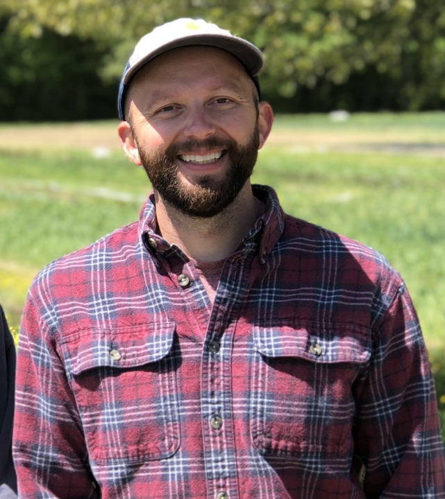 Associate Professor Joshua Puzey, smiling on a sunny day, in a plaid shirt and baseball cap.