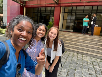 A person gives a thumb-up to the camera while taking a selfie with two others