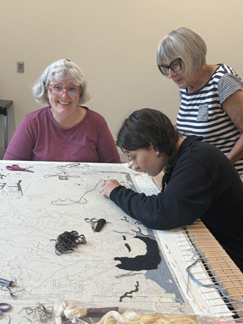 A person stitches a large canvas while two others look on.