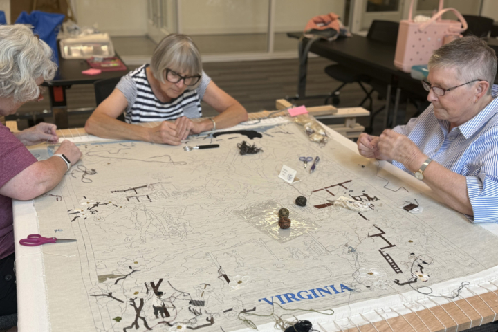 Three people stitch a large canvas that says Virginia at the bottom.