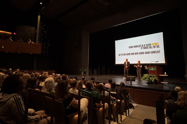 Two people stand on a stage in and auditorium near a screen that says conflict is a team sport.