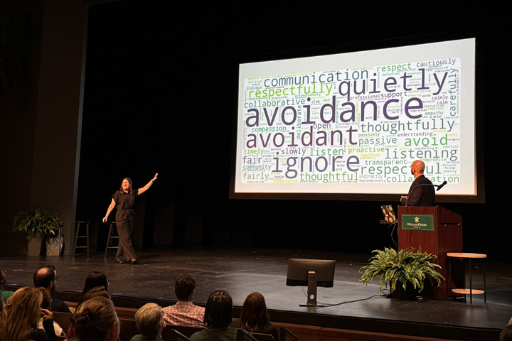 Two people stand on a stage, and one points toward a screen with a word cloud on it.