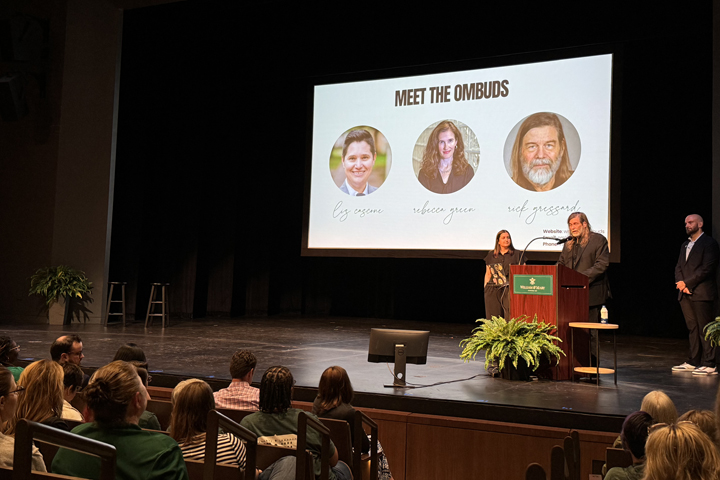A person speaks as a podium while standing near a screen that shows three people and says "Meet the ombuds."