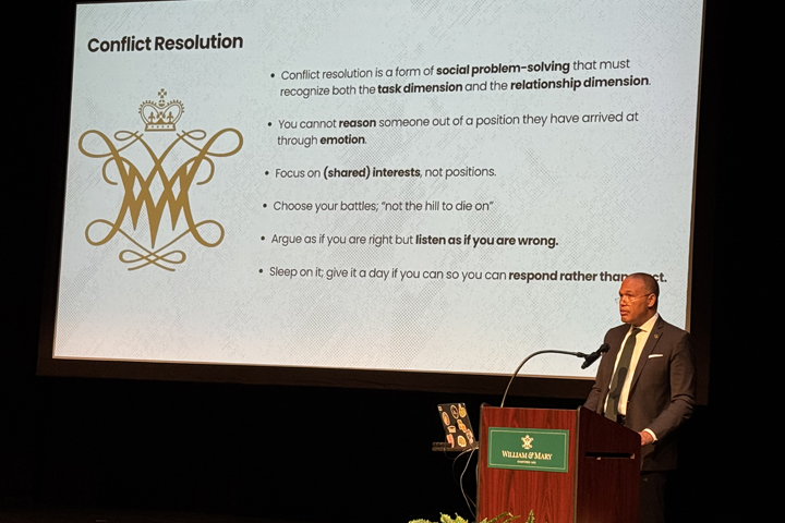 A person speaks at a podium while standing near a screen that shows several bullets about conflict resolution.