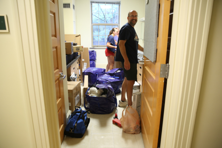 Two people stand in a dorm room with bags on the ground around their feet.