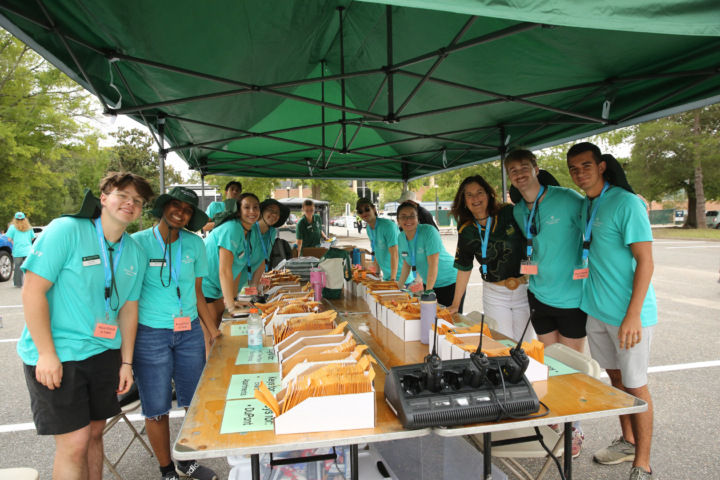 People stand under a tent near two long tables with radios, envelopes and other items lined up neatly.