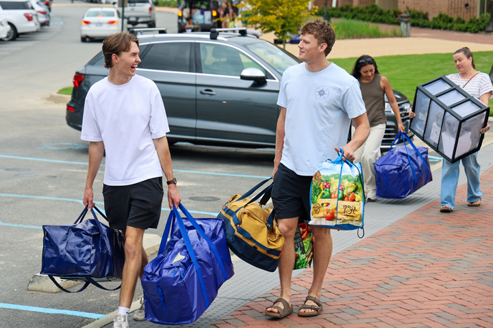 Four people carrying bags and other goods walk along a brick pathway.