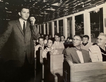 A black and white photo shows a person in a suit speaking into a microphone next to rows of people.