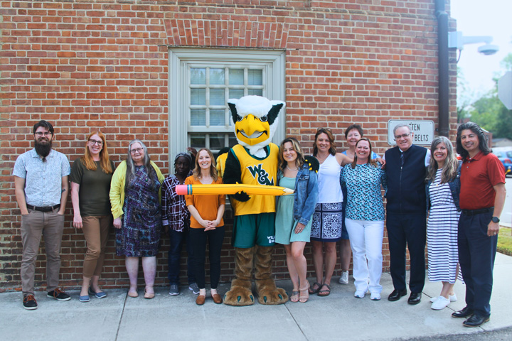 A person in a Griffin mascot holds an inflatable pencil and poses for a photo with people next to a brick building.