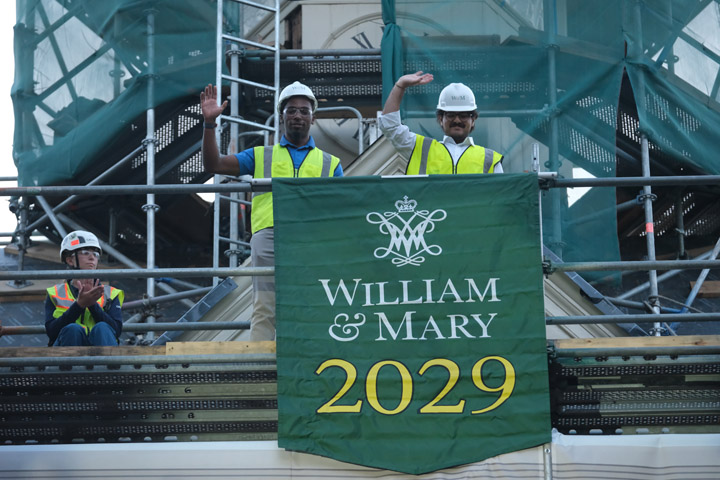 Two people in hard hats and yellow safety vests wave from atop scaffolding near a banner that says, "William & Mary 2029."