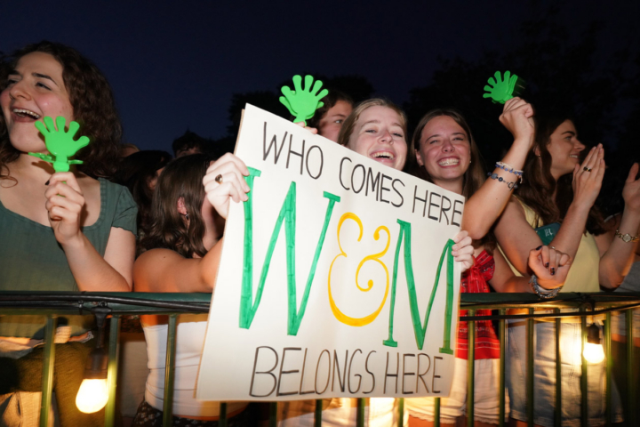 A person holds a sign that says, "Who comes here, belongs here" while standing by others who hold green, plastic hands.