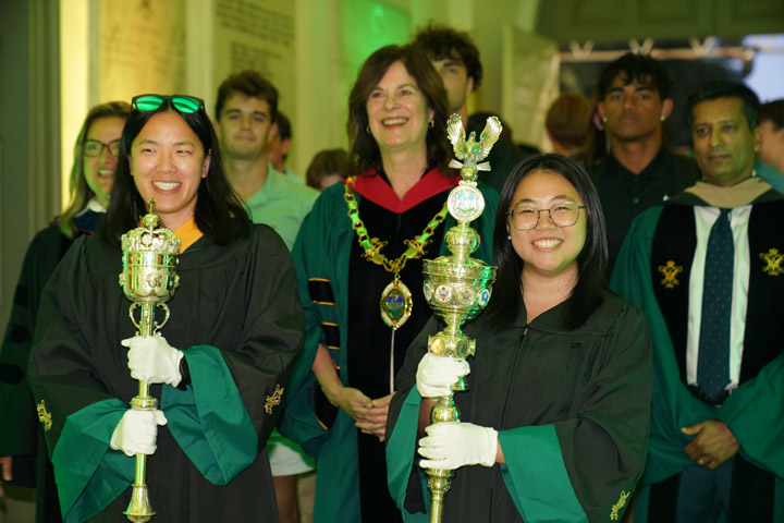 People wearing academic regalia and holding maces walk through a hallway.