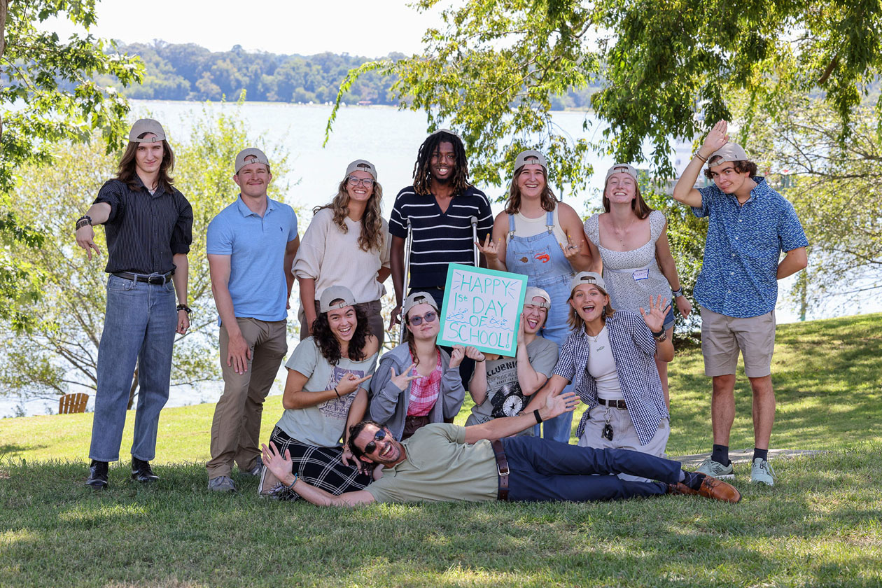 A group of people pose for a photo together near a body of water while holding a 1st day of school sign