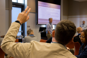 A person raises their hand while someone speaks next to a screen.