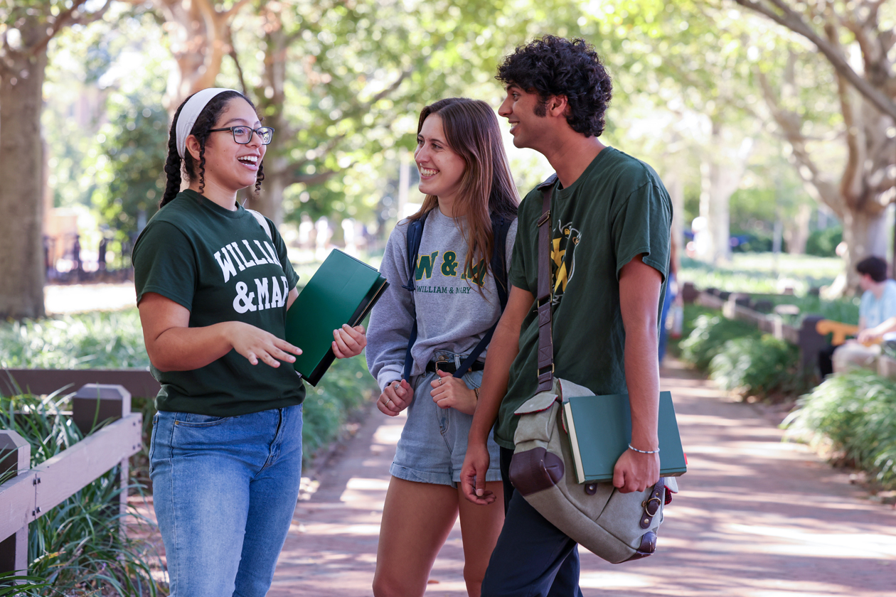 Three people wearing backpacks stand on a brick pathway and talk while holding books.