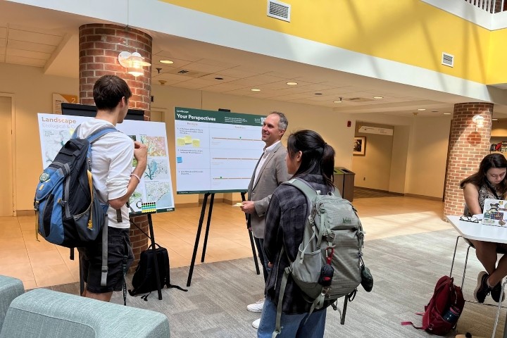 Two people wearing backpacks talk to another person who stands near two large posters.