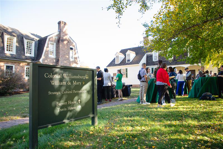 A sign saying "Colonial Williamsburg and William & Mary" stands in the grass near where people are standing by tables.
