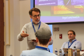 A person gestures while standing at the front of a room and instructing people sitting at table.