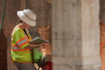 A person wearing a hardhat and safety vest writes on a notebook.
