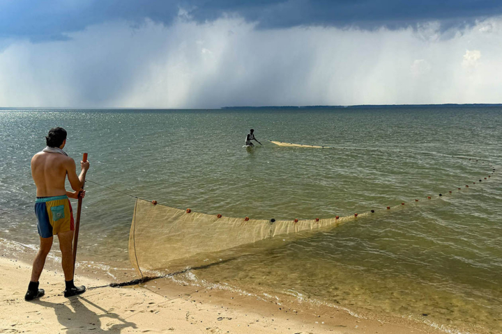 A person stands on a beach and holds one end of a net while a person in the water holds the other end.