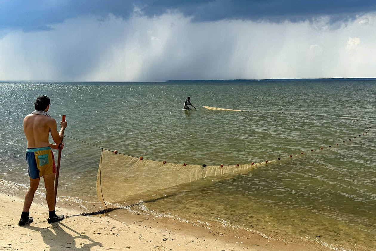 A person stands on a beach and holds one end of a net while a person in the water holds the other end.