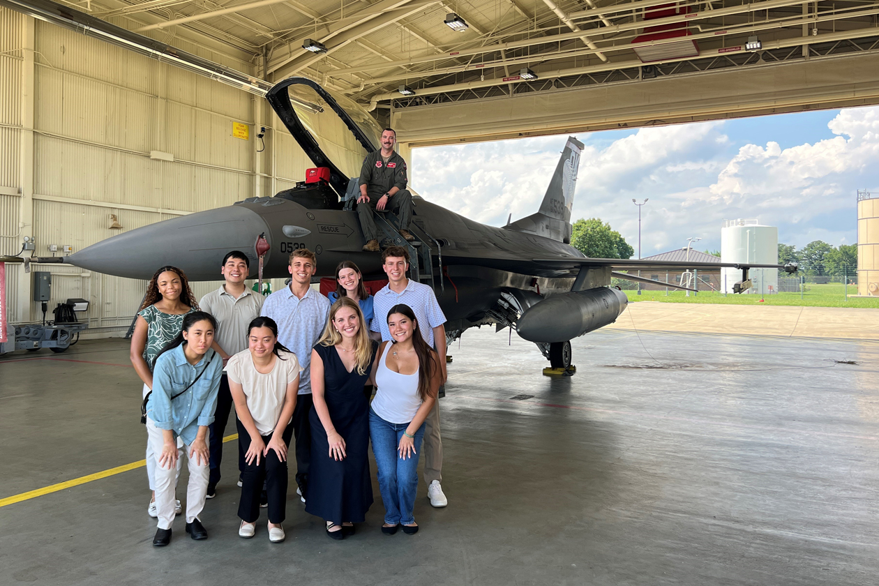 Eight people gather for a photo in front of an F-16 airplane, with a pilot sitting on top of it.