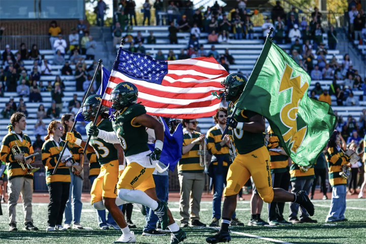 Football players run onto a field while holding flags.
