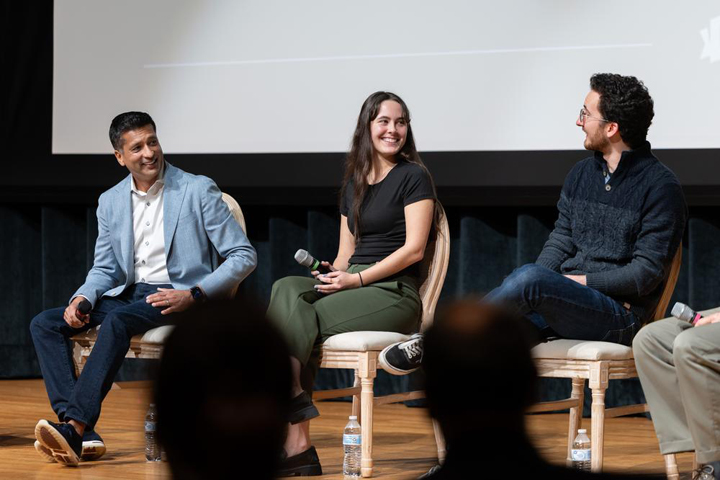 Three people smile while sitting on chairs on a stage.