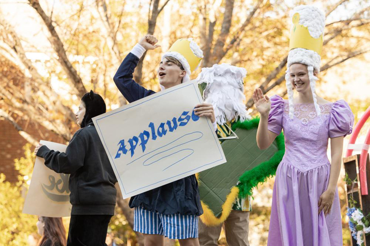 People dressed as royalty wave and hold an applause sign.