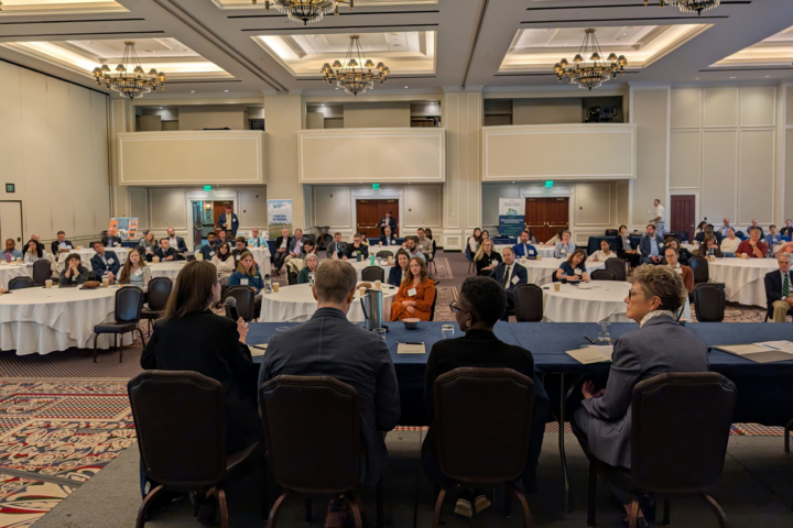 Four people sit at a long table while others sitting at tables throughout a large room listen.
