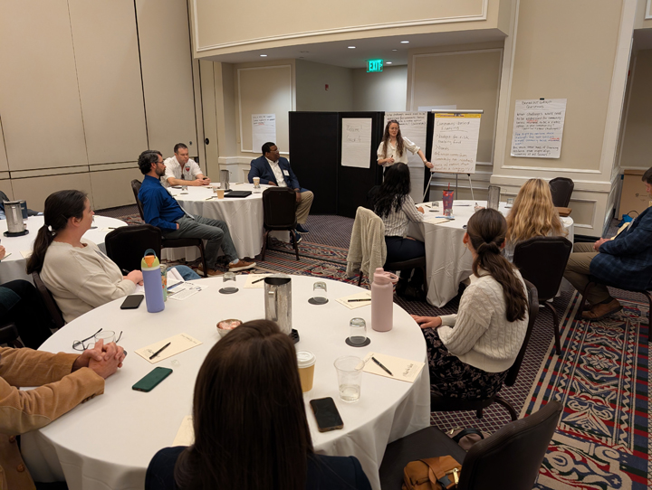 A person speaks and points at a large poster board while standing at the front of a room full of people.