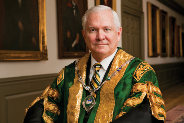 Robert Gates sits for a photo while wearing academic regalia in green and gold.