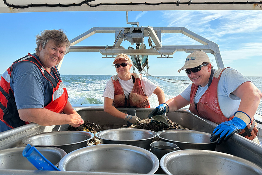 Three people wearing safety vests stand by steel buckets on a boat.