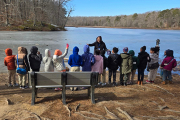 A group of children listen to an adult as they stand next to a frozen body of water at a park.