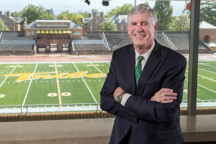 Terry Driscoll stands with arms crossed while by a window overlooking a football field.