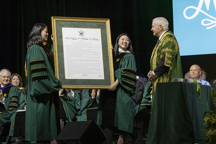 Two people hold a large, framed document and present it to Robert Gates.