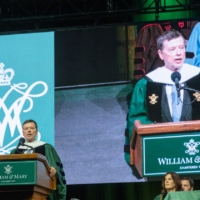 A person speaks at a podium while his image is displayed on a large screen.