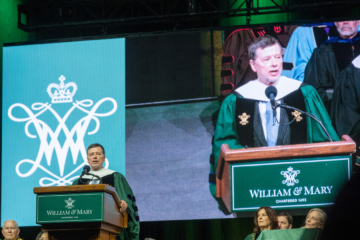 A person speaks at a podium while his image is displayed on a large screen.