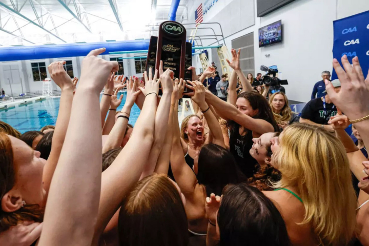 A person yells while holding up a trophy next to a pool, and a group of people with hands raised surround her.