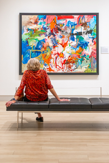 A person sits on a bench and looks at an abstract painting.