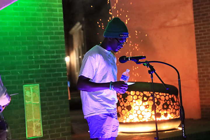 A person holds a water bottle and speaks into a microphone next to a fire.
