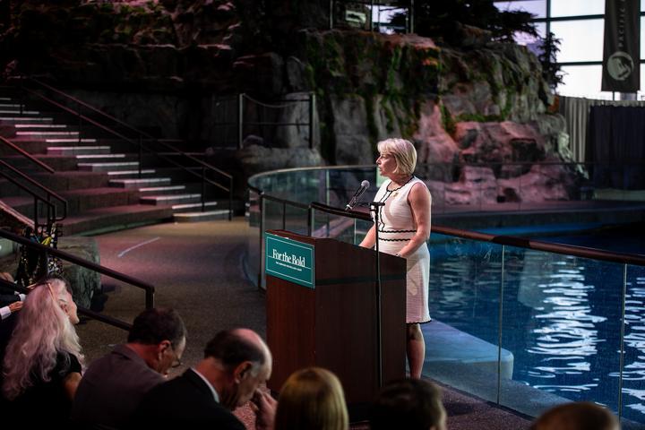 Sue Gerdelman speaks at a podium next to a large indoor pool.