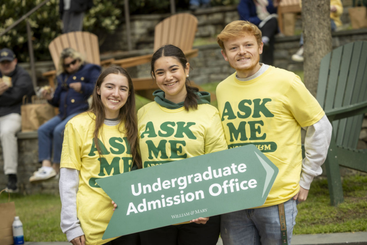 Three people in yellow shirts that say "Ask Me" hold a sign for undergraduate admission office.