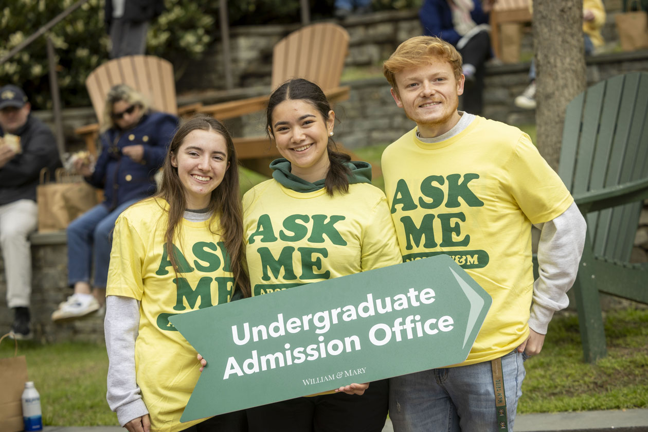 Three people in yellow shirts that say "Ask Me" hold a sign for undergraduate admission office.