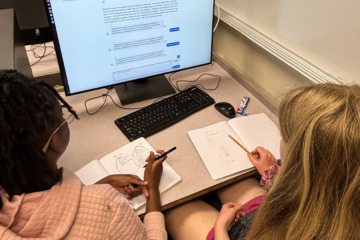 Two people sit at a desk with pens and paper and look at a computer screen.