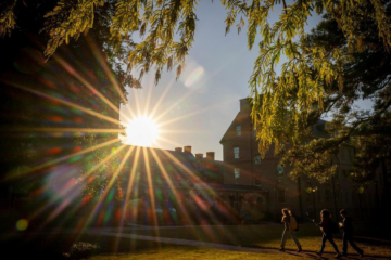 People walk on a brick path toward a brick building while the sun shines brightly above.