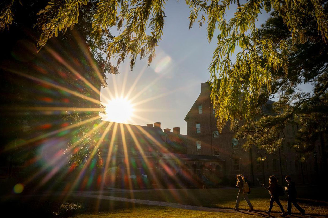 People walk on a brick path toward a brick building while the sun shines brightly above.